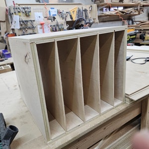 May include: A wooden organizer with four compartments, made of light-colored plywood. The organizer is sitting on a wooden workbench.