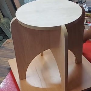 May include: A light-colored wooden stool with a round top and four curved legs. The stool is made of light-colored wood and has a simple, modern design. The stool is approximately 18 inches tall.