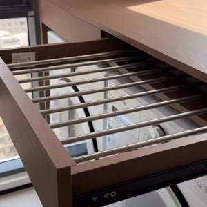 May include: A wooden clothes drying rack with stainless steel bars, partially pulled out from a brown cabinet. The rack is designed for air-drying garments. The cabinet is near a window, suggesting an indoor laundry setup.