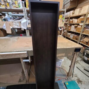 May include: A tall, dark brown wooden shelf unit with a rectangular shape. The shelf is empty and stands upright on a dark gray mat. The unit is in a workshop setting, with shelves and tools visible in the background.