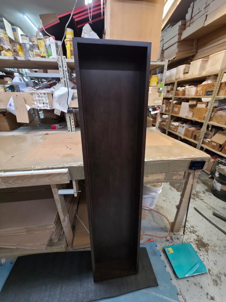 May include: A tall, dark brown wooden shelf unit with a rectangular shape. The shelf is empty and stands upright on a dark gray mat. The unit is in a workshop setting, with shelves and tools visible in the background.