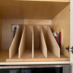 May include: A wooden cabinet organizer with five vertical dividers for storing cutting boards. The organizer is installed in a kitchen cabinet above a stainless steel oven.