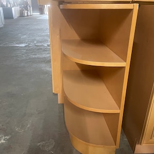 May include: A light wood corner cabinet with three shelves. The shelves are curved to fit the corner and are open on all sides. The cabinet is made of a light wood with a smooth finish.