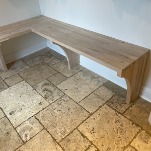 May include: A corner bench made of light wood with a natural finish. The bench is mounted to the wall and has a simple design. The bench is in a room with a tiled floor.