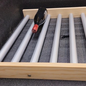 May include: A wine rack featuring two light-colored wooden side rails connected by six parallel grey cylindrical rods. A single dark glass wine bottle with a red foil cap rests horizontally between two of the rods on the rack structure.