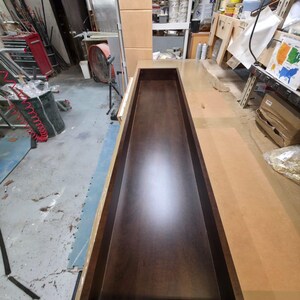 May include: A rectangular, dark brown wooden box with a recessed interior. The box is open at the top and appears to be part of a larger woodworking project, possibly a cabinet or drawer. The wood has a polished finish.