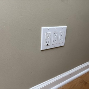May include: A white electrical outlet cover with three standard electrical outlets is mounted on a beige wall. The wall has a slightly textured surface. The bottom of the image shows a section of a white baseboard and hardwood flooring.