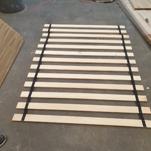 May include: A wooden slat bed frame with black straps. The slats are made of natural wood and are evenly spaced. The straps are attached to the slats and are used to support the mattress.