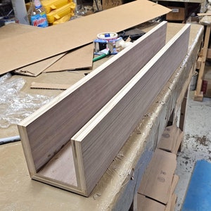 May include: A wooden shelf unit with a U-shaped opening, made from two pieces of wood joined together. The wood has a light brown color with darker brown grain patterns.