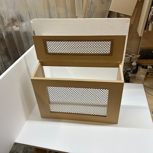 May include: A wooden radiator cover with a decorative metal grille. The cover is open, revealing the interior. The cover is a light brown color.