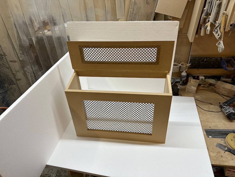 May include: A wooden radiator cover with a decorative metal grille. The cover is open, revealing the interior. The cover is a light brown color.