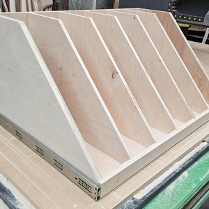 May include: A wooden storage shelf with five compartments, each with a slanted front. The shelf is made of light-colored wood and has a silver metal drawer slide on the bottom.