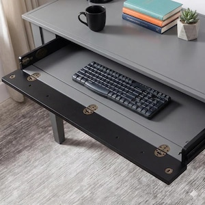 May include: A gray desk with a fold-down front panel featuring brass-colored hinges. A black computer keyboard sits on the extended tray. On the desk surface, there is a black mug, a stack of three books, and a small succulent in a geometric pot.