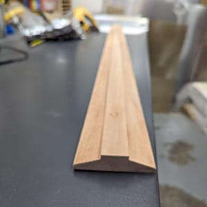 May include: A close-up of a wooden trim piece with a beveled edge and a raised center section. The trim is light brown in color and has a smooth finish.