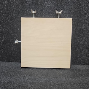 May include: A square piece of light wood with two silver wing nuts attached to the top. The wood is sitting on a black textured surface.