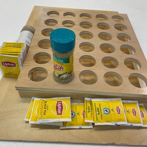 May include: A wooden spice rack with 24 round holes, a teal spice jar labeled "Mrs. Dash", a stack of yellow tea bags labeled "Lipton Black Tea", and a brown spice bottle.