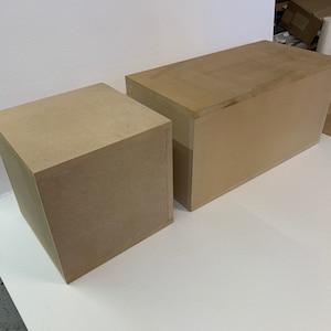 May include: Two unfinished, rectangular wooden boxes of different sizes. The boxes are made of light brown wood and have a simple, boxy design. They are placed on a white surface, with a white wall in the background. The boxes appear to be ready for finishing or use.