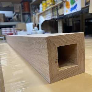 May include: A long, rectangular piece of wood with a square hole cut out of the center. The wood is light brown and has a natural grain pattern.
