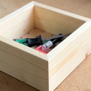 May include: A wooden box with a light wood finish. The box is empty and has a few small holes in the bottom. The box is sitting on a brown surface.