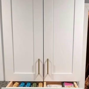 May include: White kitchen cabinets with gold handles and a pull-out drawer containing coffee pods.