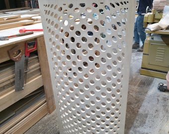 May include: A tall, unfinished wooden cabinet with a series of evenly spaced, circular cutouts. The cabinet has a rectangular frame and a flat top. The design suggests a modern, minimalist aesthetic. The cabinet is in a workshop setting.