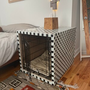 May include: A dog crate with a black and white checkered design. The crate has a metal door and a beige cushion inside. The crate is placed on a patterned rug and is next to a bed.