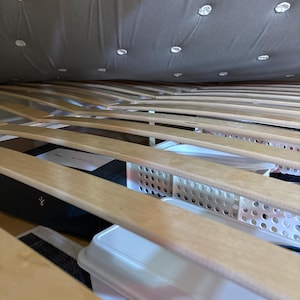 May include: A wooden slat bed frame with a gray quilted mattress. The bed frame has storage space underneath, which is filled with white plastic storage containers.