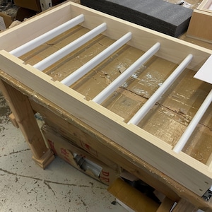 Puede incluir: Un cajón de madera con clavijas blancas para organizar objetos. El cajón está hecho de madera clara y tiene un acabado natural. Las clavijas están espaciadas uniformemente y están diseñadas para mantener los objetos en su lugar.