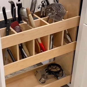 May include: A pull-out kitchen cabinet organizer with various cooking utensils. The organizer is made of light wood and features multiple compartments. Utensils include knives, spatulas, whisks, and other kitchen tools. The cabinet is white.