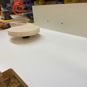May include: A woodworking scene featuring a white tabletop with a wooden, circular platform attached to a black base. A white rectangular piece of wood is in the background. Power tools are visible in the background.