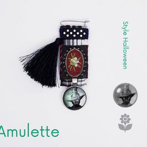 May include: A black and white fabric brooch with a black tassel and a round glass charm featuring a black cat wearing a witch's hat. The charm is attached to a silver pendant. The brooch is labelled "Style Halloween".