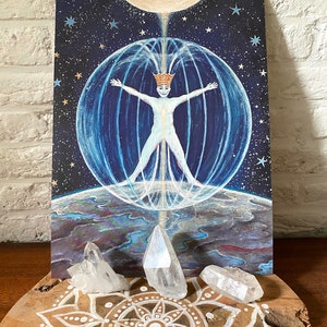 May include: A painting of a person with outstretched arms inside a sphere of light, surrounded by stars and a crescent moon. The person is wearing a crown and is surrounded by a blue and white aura. The painting is on a wooden surface with a mandala design and three clear quartz crystals.