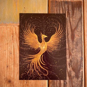 Puede incluir: Una impresión de un fénix dorado con las alas extendidas sobre un fondo marrón oscuro. El fénix está detallado con tonos dorados y amarillos, y el fondo está salpicado de pequeños puntos dorados. La obra de arte se muestra sobre una superficie de madera.