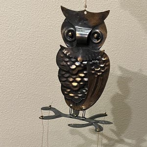 May include: A metal owl wind chime with a brown and copper finish. The owl is perched on a branch with three hanging metal leaves.