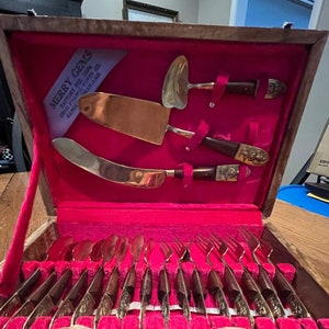 May include: A wooden box with a red velvet interior containing a set of 12 gold-toned flatware pieces. The box has a label that reads "Merry Gems Factory Try Them One Each You Will Be Delighted."