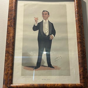 May include: A framed Vanity Fair print featuring a full-length caricature of M. John Hare in formal attire. The print is in a decorative wooden frame with a burl wood finish. The print is signed "Spy".