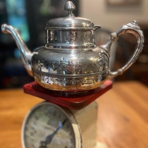May include: A polished silver teapot featuring an ornate handle, a curved spout, and a lid with a small finial. The body is decorated with engraved floral patterns and sits atop a red platform on a mechanical scale with a circular dial.
