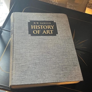 May include: A hardcover book titled "H.W. Janson History of Art" with a gray fabric cover. The title is in gold lettering on a black rectangular label. The book is resting on a dark surface.