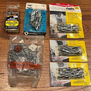 May include: Assortment of aluminum rivets in various packaging. The rivets are silver and come in different sizes. The packaging includes a clear plastic bag, a yellow and black package with the text "Swingline Aluminum Rivets", and a blue and white package with the text "Pop Rivets".