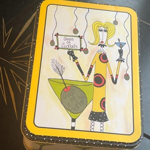 May include: A rectangular metal tin featuring a yellow border with white dots. The lid displays a cartoon illustration of a blonde figure holding a martini glass and a sign that reads Queen of Cocktails, with a large green martini glass below.