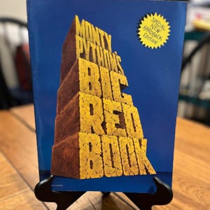May include: A blue hardback book titled Monty Python's Big Red Book with large, textured yellow block lettering. A yellow starburst graphic in the top right corner contains the text Special New Hardback Edition in black capital letters.