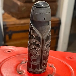 May include: A dark brown, cylindrical carved figurine featuring a stylized head with closed eyes and a small mouth. The body is decorated with intricate, light-colored etched floral patterns and vertical lines, resembling a robe or garment.