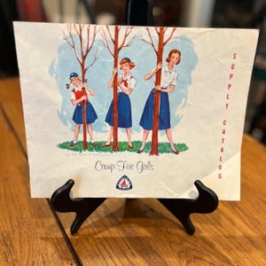 May include: Vintage Camp Fire Girls Supply Catalog cover. The illustration features three girls in blue skirts and white blouses, holding onto trees. The text "Camp Fire Girls" and "Supply Catalog" are visible. The background is a light blue sky.