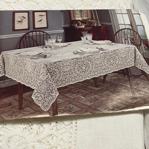 May include: A beige lace tablecloth covers a wooden dining table set for a meal. The tablecloth has a detailed floral pattern and scalloped edges. The table is set with plates, silverware, and glassware. Two framed pictures hang on the wall.