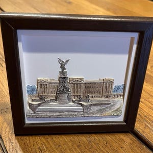 May include: A framed relief sculpture depicting Buckingham Palace. The sculpture features a detailed facade of the palace, a monument, and surrounding landscaping. The frame is dark brown, and the background is white.