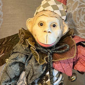 May include: A whimsical monkey figurine dressed as a jester. The monkey has a cream-colored face with a wide smile and blue eyes. It wears a checkered hat, a ruffled collar, and a patterned jacket with bells. The jester's outfit is a mix of textures and colors.
