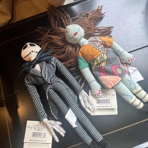 以下が含まれることがあります： 刺繍された顔のディテールを持つ、ジャックとサリーの2体のぬいぐるみ。ジャックはコウモリの蝶ネクタイが付いた黒と白のピンストライプのスーツを着ています。サリーは毛糸の髪とパッチワークのドレスを身に着けています。各人形にはMini Bean Bagと書かれた白いタグが付いています。