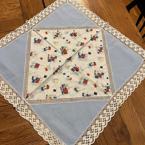 May include: Square, vintage-style tablecloth with a light blue border and a central patterned square. The center features a white background with red, blue, and yellow geometric shapes. The border is trimmed with delicate white lace.