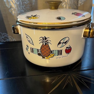 May include: White enamel pot with lid, featuring a gold rim and handle accents. The pot is decorated with colorful fruit and leaf illustrations, including a pineapple and strawberry. The text "Georges Briard" is printed on the pot.
