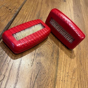 May include: Two red, rectangular carpet sweepers with natural-colored bristles. The sweepers are made of a glossy red plastic and have a ridged design on the top. The bristles are visible in a rectangular opening.